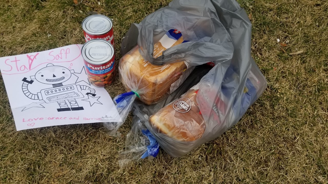 photo of typical grocery delivery with two loaves of bread and two cans of food. Delivery also includes a handwritten note by a child that reads, "stay saff'