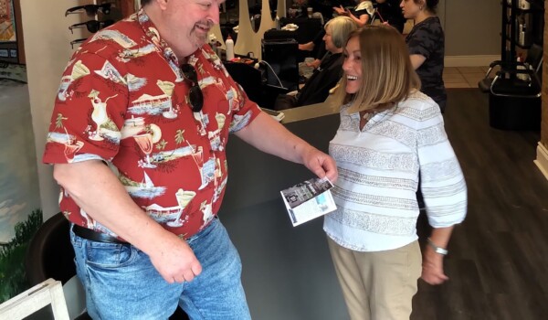 A man in a red shirt and blue jeans gives a flyer to a persn wearing a white and grey shirt and beige pants.